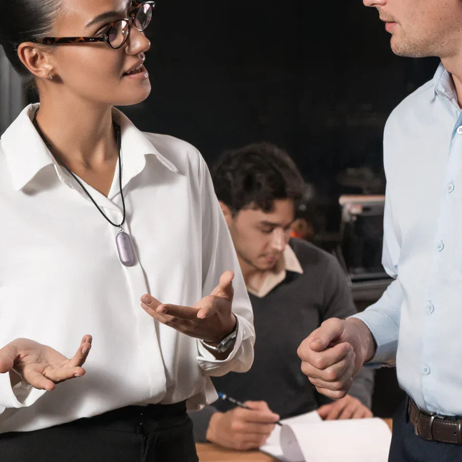 A woman wearing a purple Plaud NotePin on a lanyard while talking to a colleague in an office.
