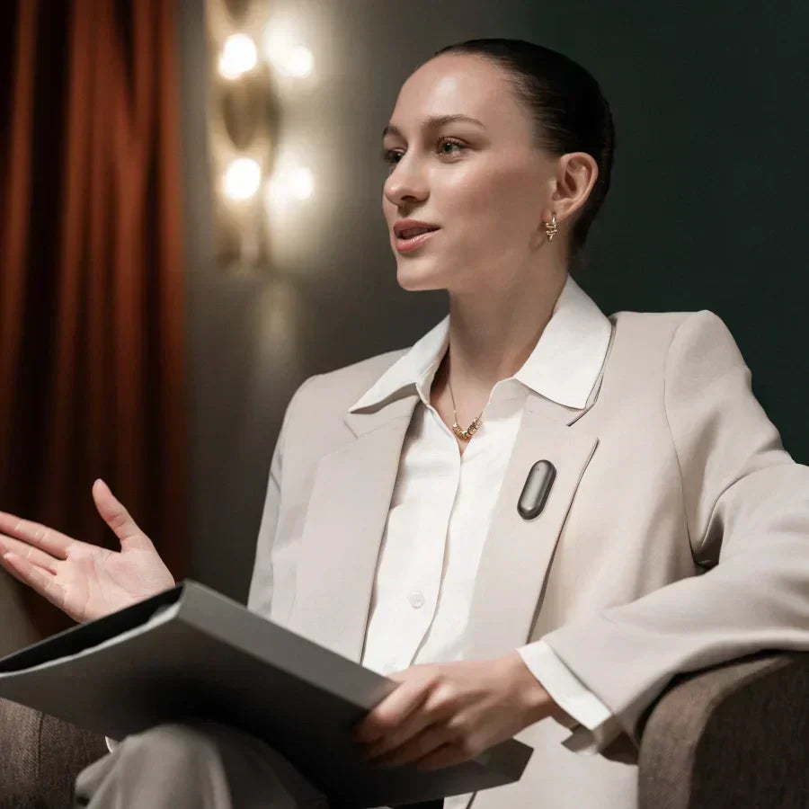 Woman in a beige blazer speaking with a gray Plaud NotePin worn on her lapel.