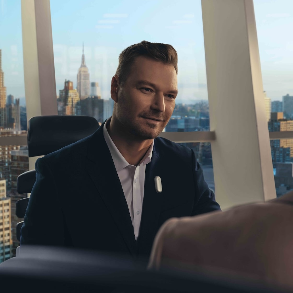 Man in a suit wearing a silver Plaud NotePin on his lapel in a high-rise office with a city view.
