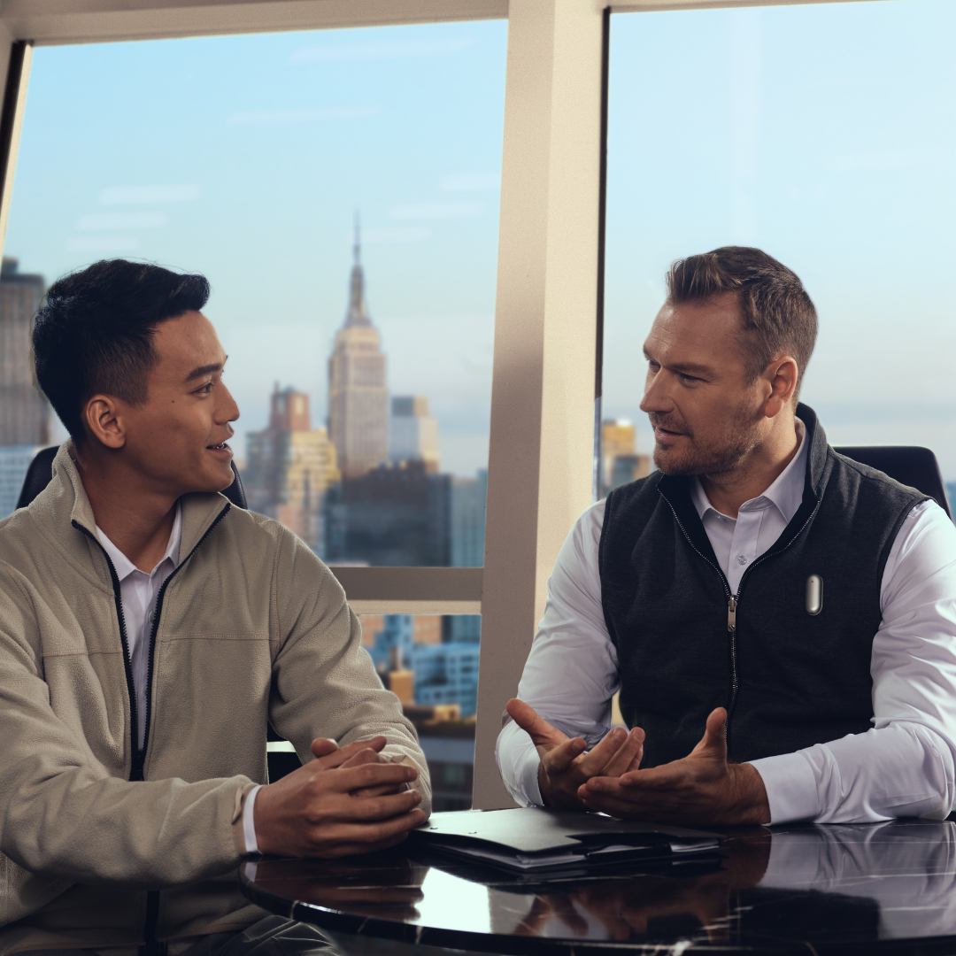 Two men in a business meeting; one wears a silver Plaud NotePin on his vest to record the conversation.
