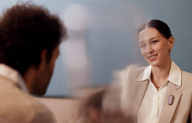 A woman wearing a purple Plaud NotePin on her blazer lapel while speaking with a man in a professional setting.