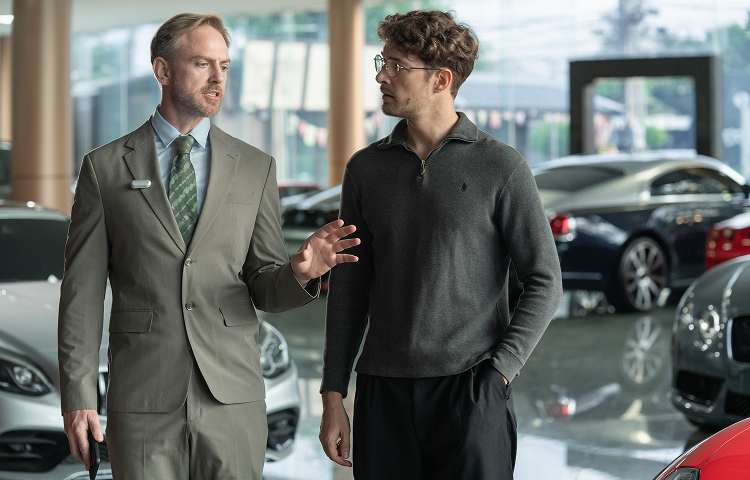 Salesman wearing a silver Plaud NotePin on his lapel talks to a customer in a car showroom.