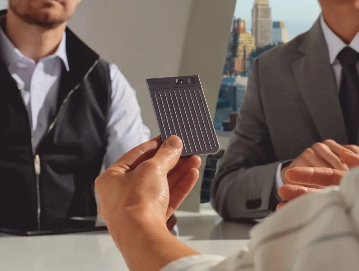 A hand holds a grey Plaud Note Pro during a business meeting with colleagues in a professional office.