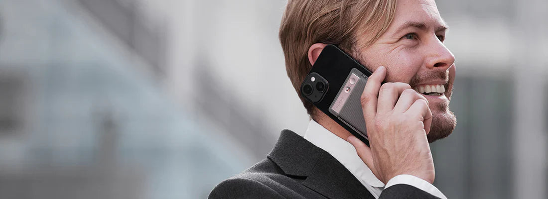 Man in a suit takes a phone call with a silver Plaud Note attached to the back of his smartphone.