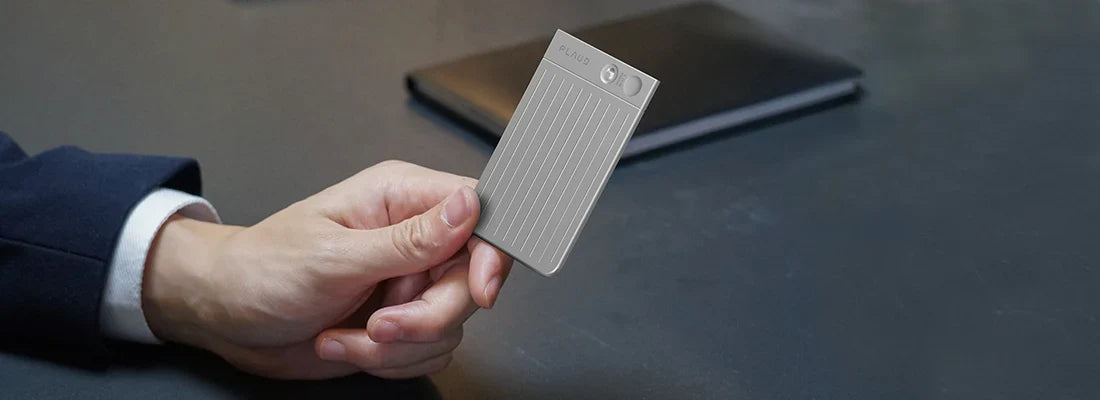 A person's hand holding a silver Plaud Note Pro over a desk with a black notebook.