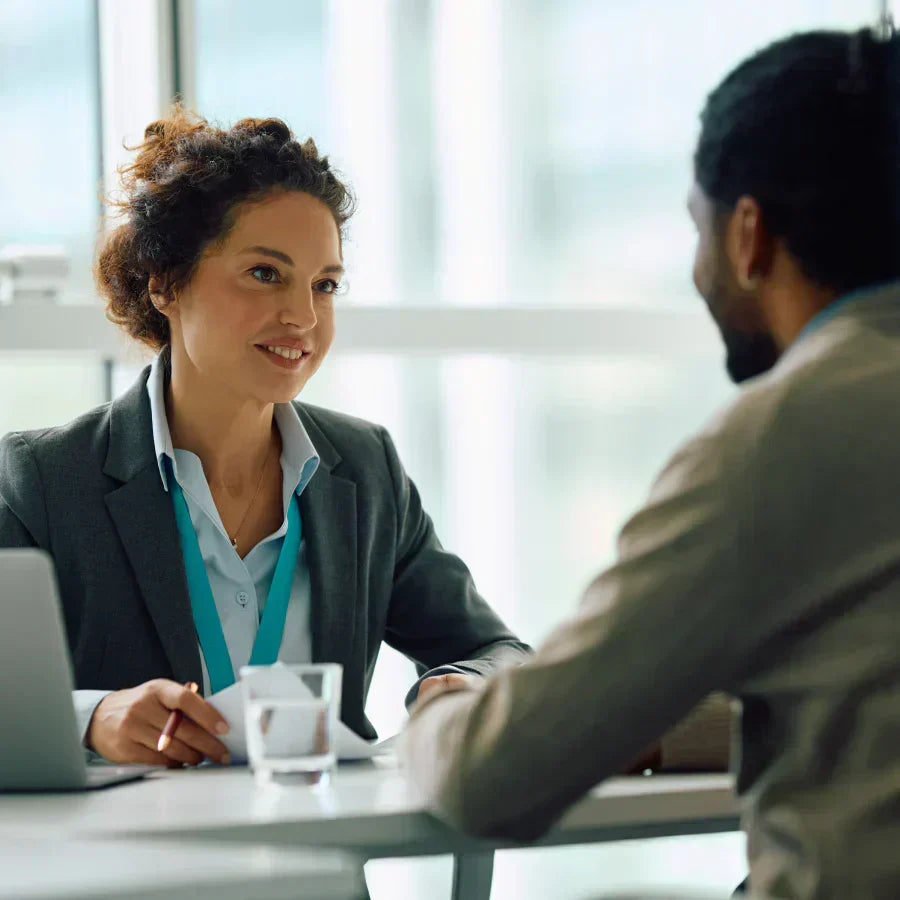 A professional woman in a grey blazer and teal lanyard conducting a business meeting or interview in a bright office.