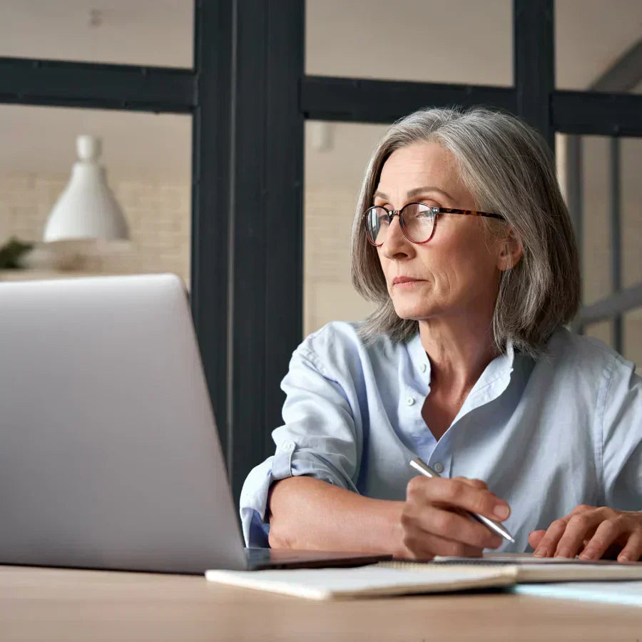 Woman with glasses looking at a laptop and taking notes at a desk.
