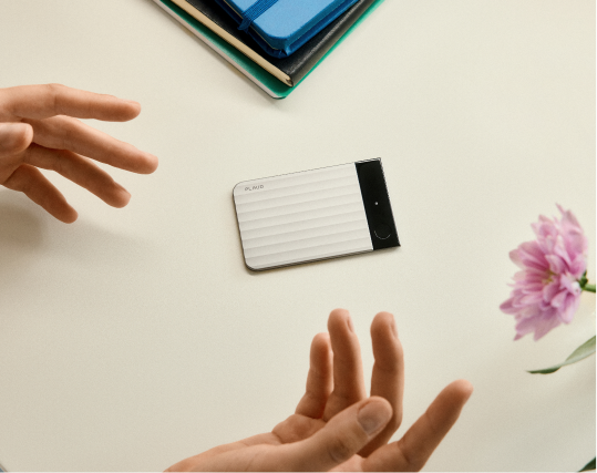 Silver Plaud Note Pro on a table between gesturing hands, next to notebooks and a pink flower.