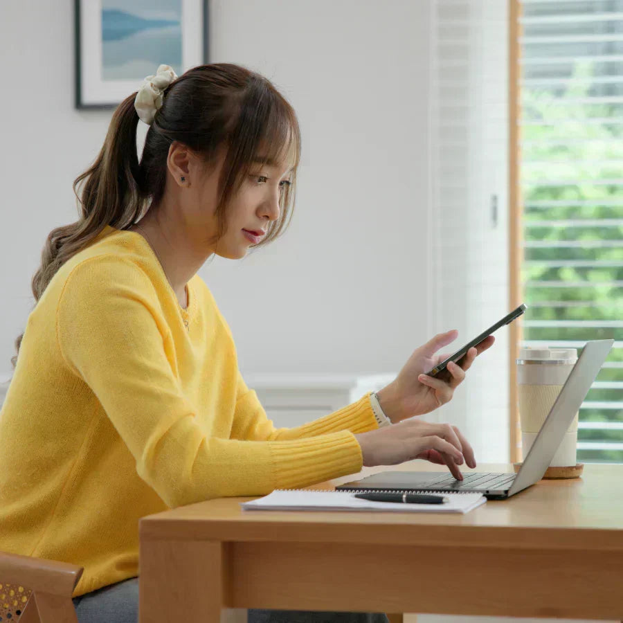 A woman in a yellow sweater uses a laptop while holding a phone with a black Plaud Note attached to the back.