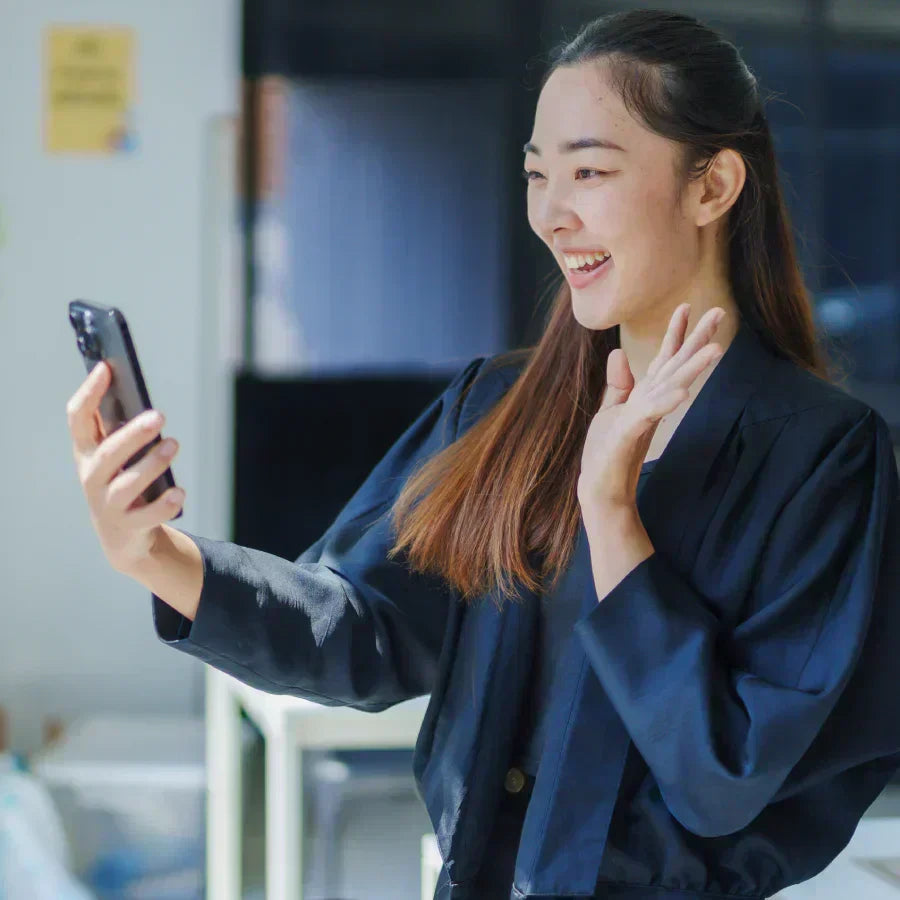 A woman waves during a video call on her phone, which has a black Plaud Note AI voice recorder attached.