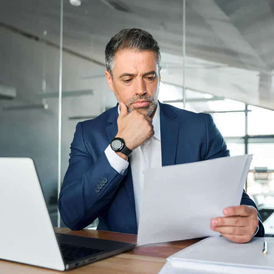 Professional in a suit reviewing documents at a desk with a laptop in an office.