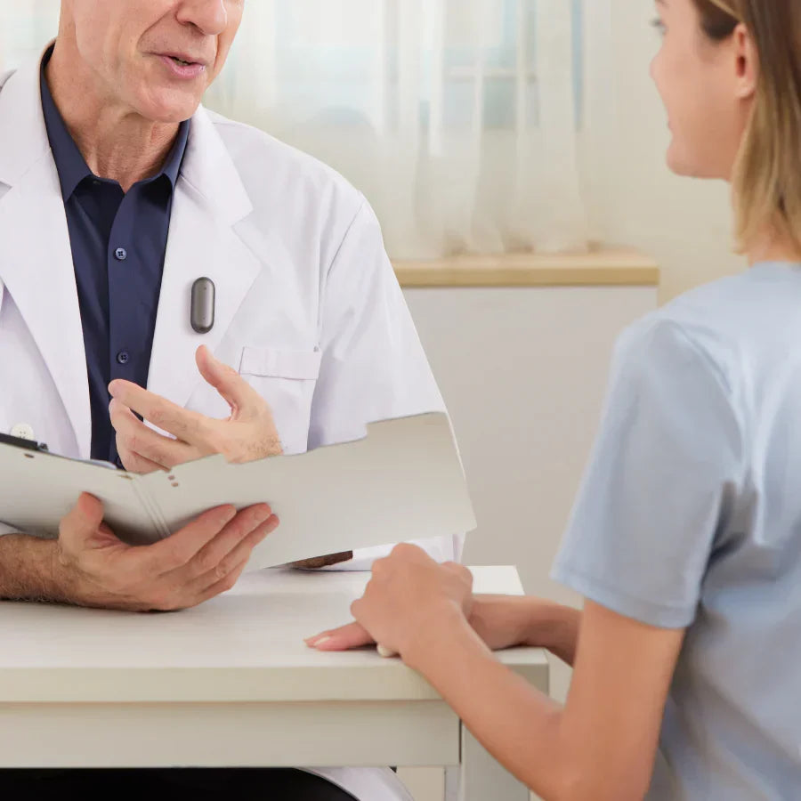 A doctor in a white coat wears a black Plaud NotePin while speaking with a patient.