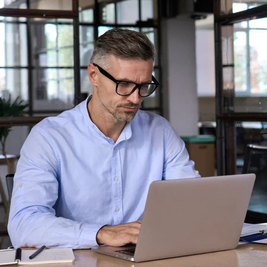 A professional man in a blue shirt and glasses working on a laptop in a modern office.