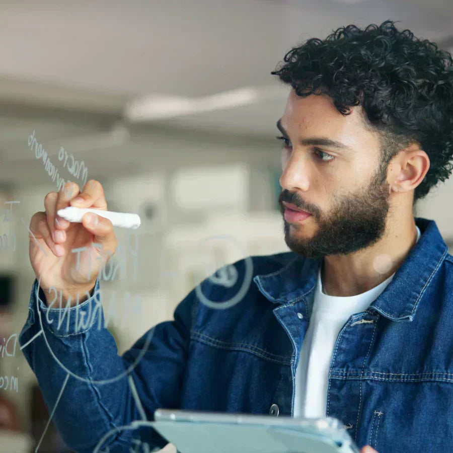Man writing on a glass board while holding a phone with a silver Plaud Note attached.