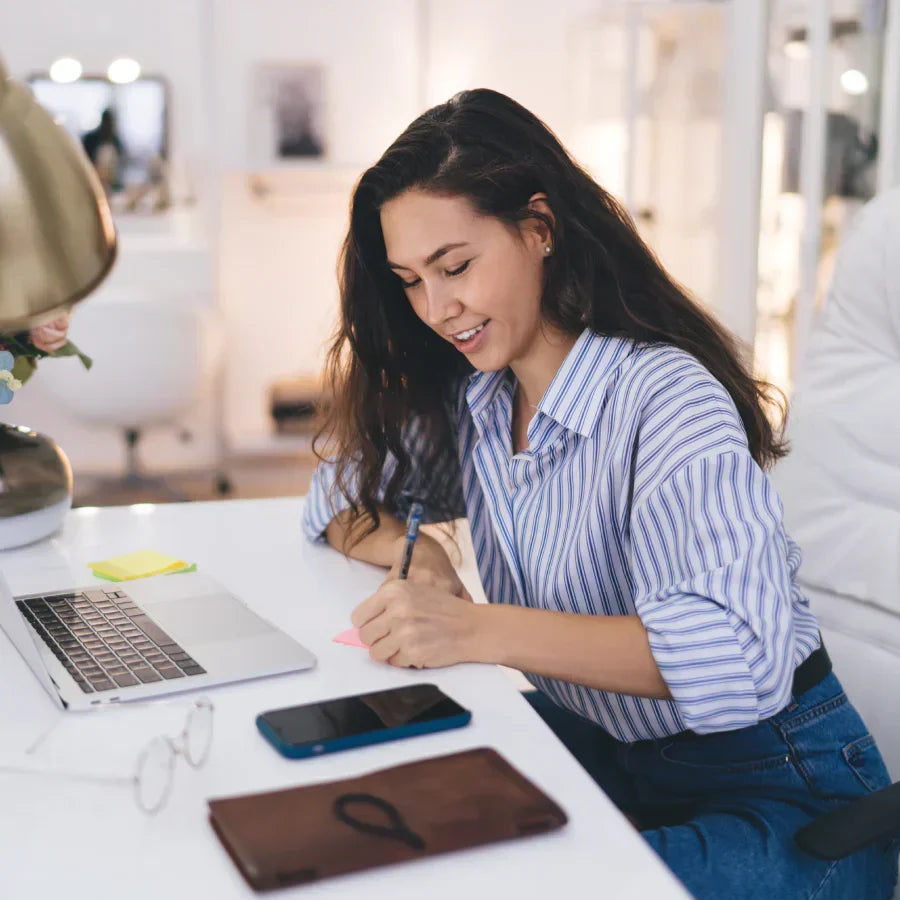 Woman writing at a desk with a black Plaud Note in a leather case, smartphone, and laptop.