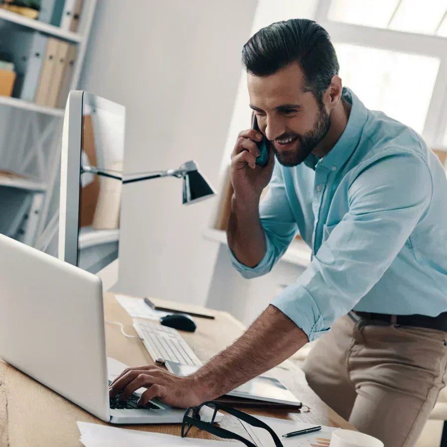 A professional talking on a smartphone with a black Plaud Note attached, recording a call while using a laptop.