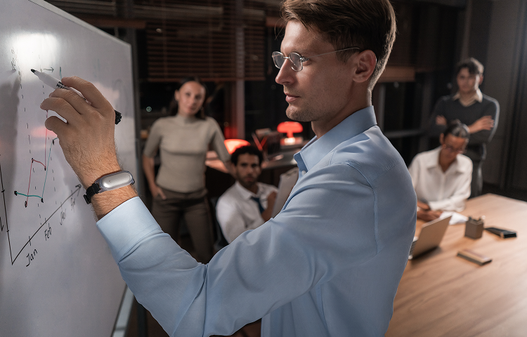Man wearing a silver Plaud NotePin on his wrist while drawing a graph on a whiteboard during a team meeting.