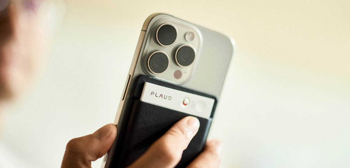 A silver Plaud Note in a black case attached to an iPhone, with a finger pressing the record button.