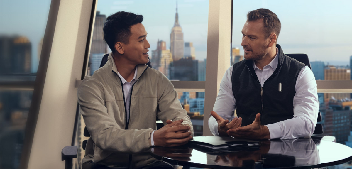 Two men talking in a high-rise office; one wears a silver Plaud NotePin on his vest to capture the conversation.