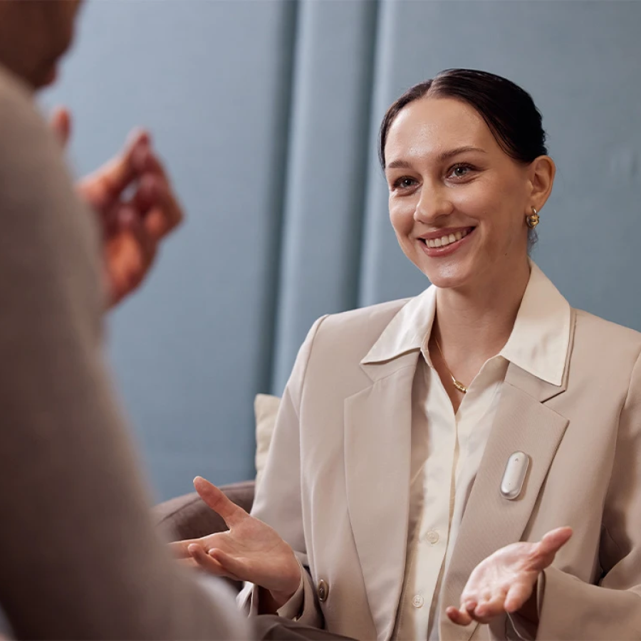 Smiling woman wearing a silver Plaud NotePin on her blazer lapel while talking to a colleague.