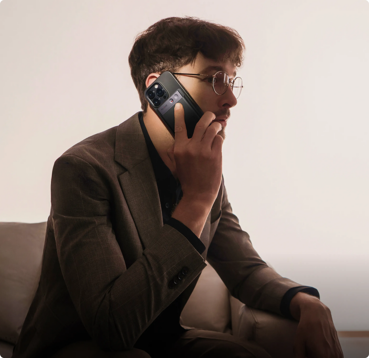 An executive in a brown suit and glasses holds a smartphone with a black Plaud Note attached to its back, talking on a call.