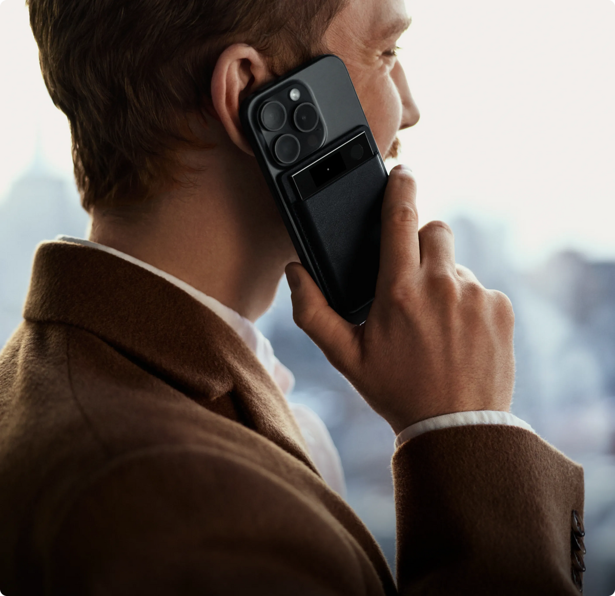 An executive in a brown coat holds a black phone with a Plaud Note Pro attached to his ear, looking out a window.