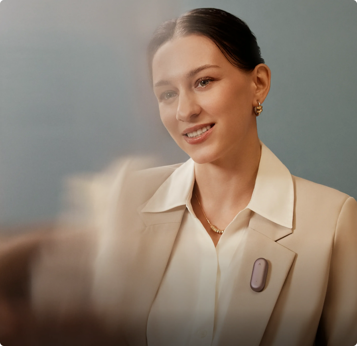 A sales in a light blazer wears a purple Plaud NotePin on her lapel against a soft blue background.