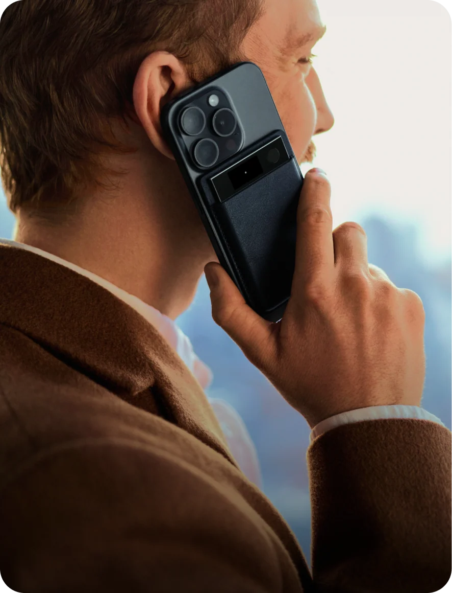 A man in a brown coat holds a black smartphone to his ear with a black Plaud Note Pro AI voice recorder attached to the back.
