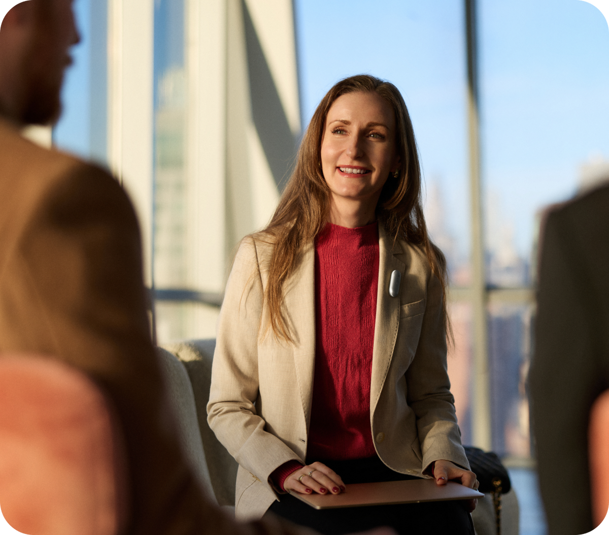 A woman in a beige blazer and red turtleneck smiles during a meeting while wearing a silver Plaud wearable AI recorder on her lapel.