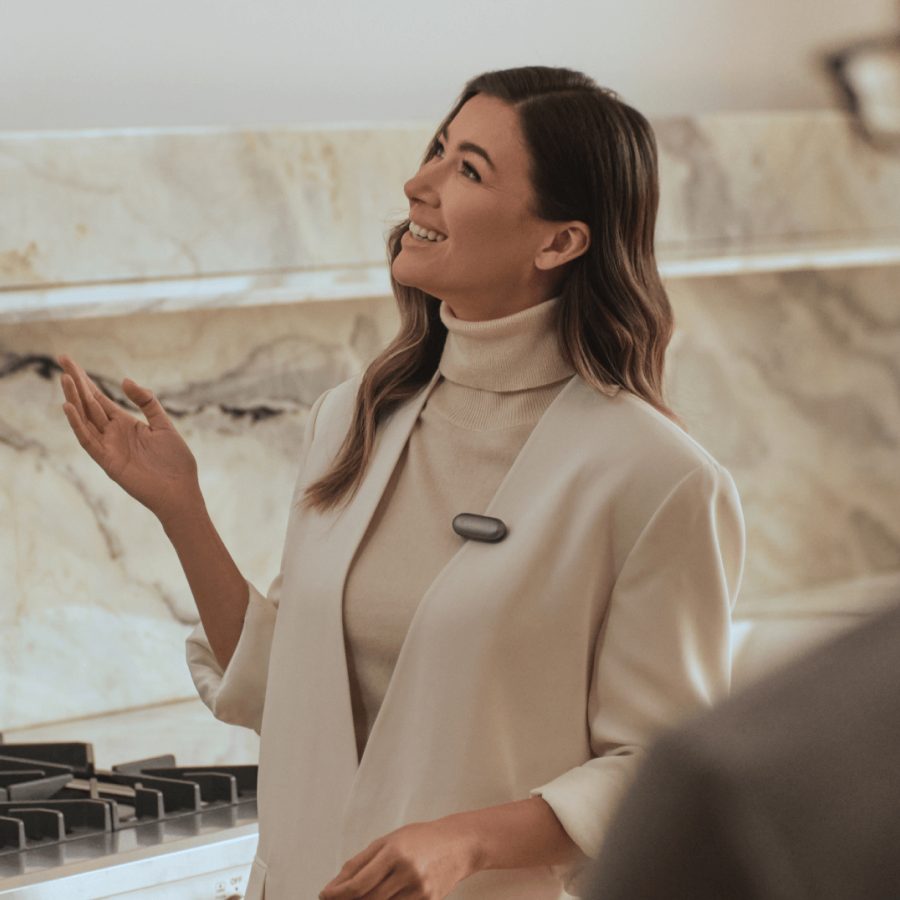 A woman wearing a gray Plaud NotePin on her lapel while gesturing and smiling in a modern kitchen.