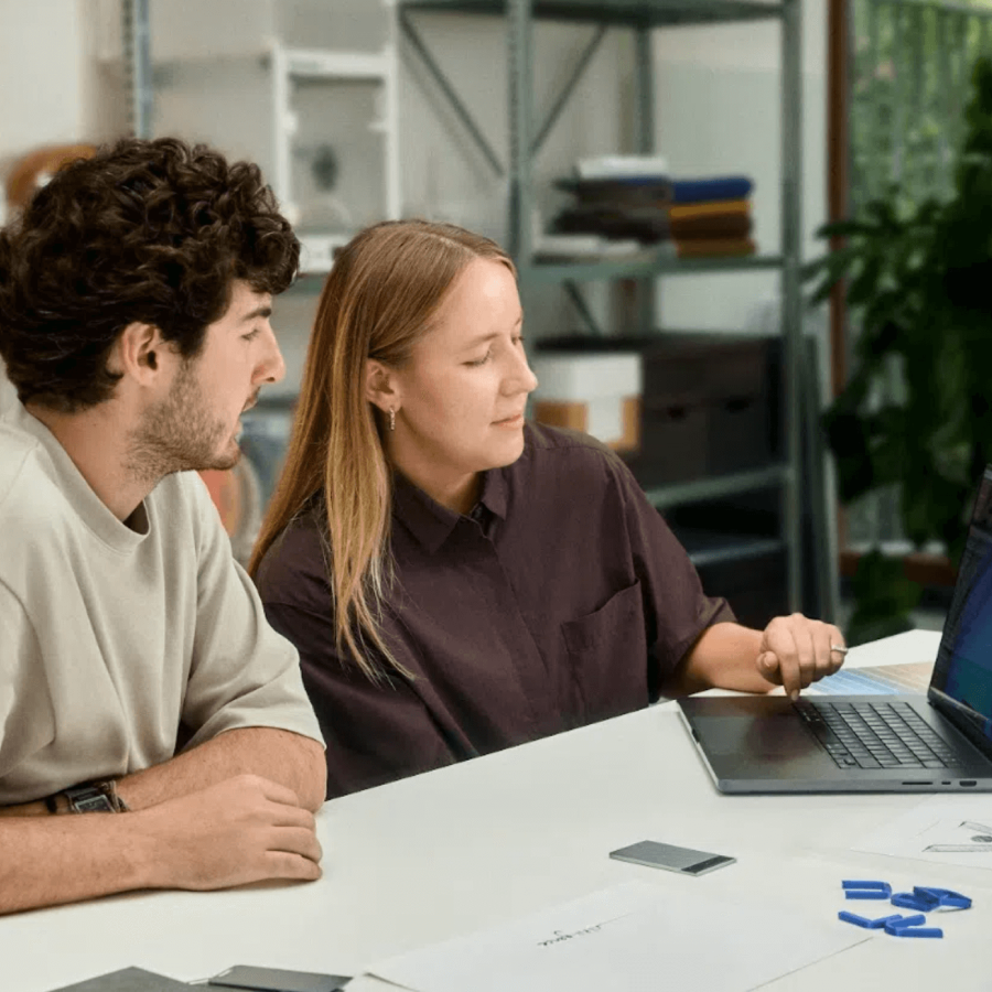 Two colleagues collaborate at a desk with a laptop and a silver Plaud Note AI voice recorder.