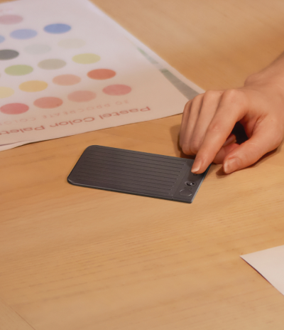 A hand operates a black Plaud Note Pro on a wooden table next to a pastel color palette.