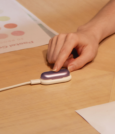 Hand touching a purple Plaud NotePin charging on a wooden desk.