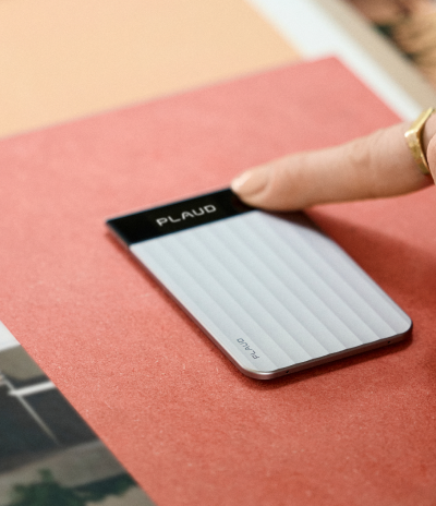 A person touches the screen of a silver, ribbed Plaud Note Pro AI voice recorder on a red background.