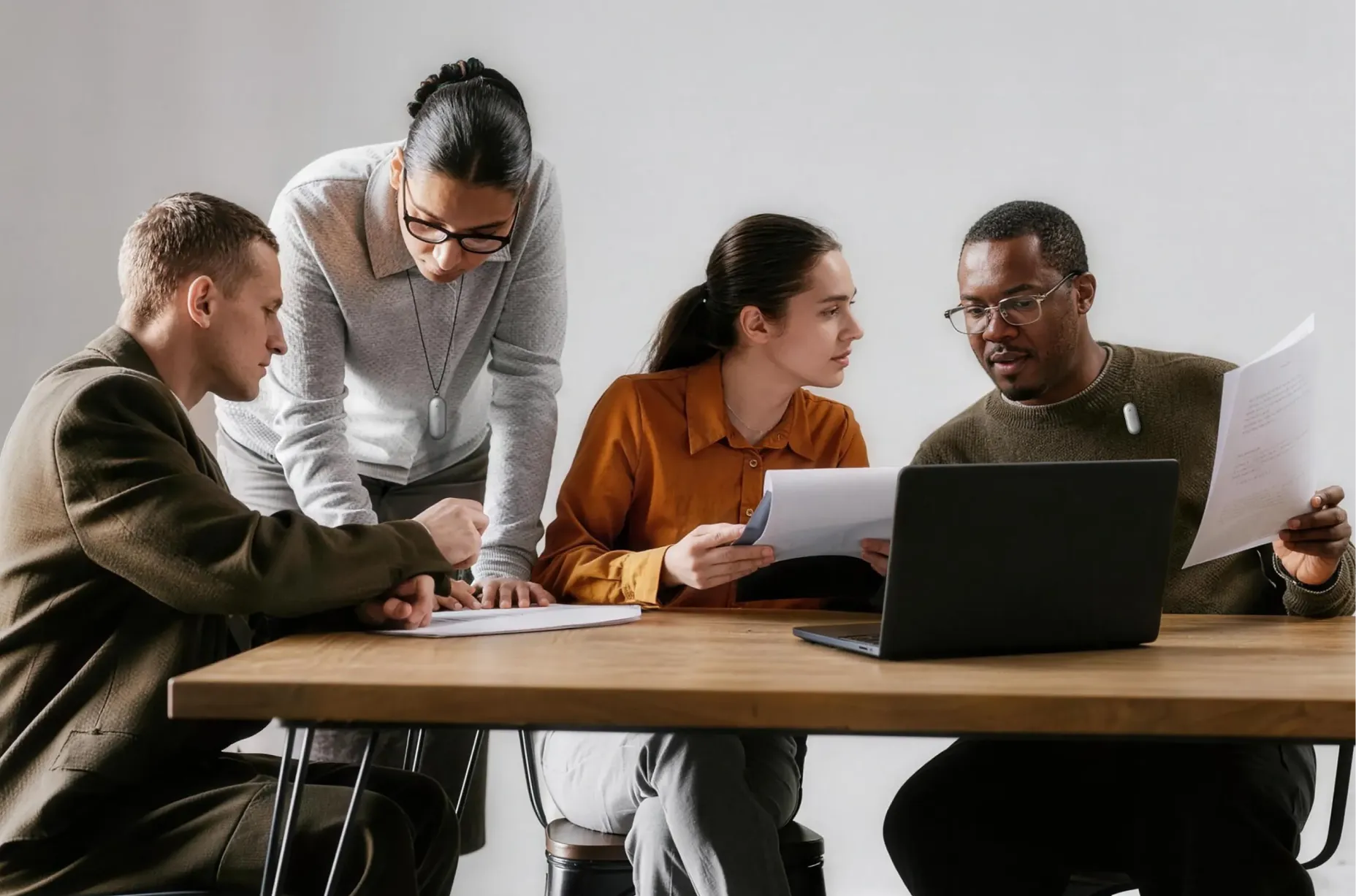 Four professionals collaborate around a wooden table with a laptop and documents, wearing silver Plaud wearable AI recorders.