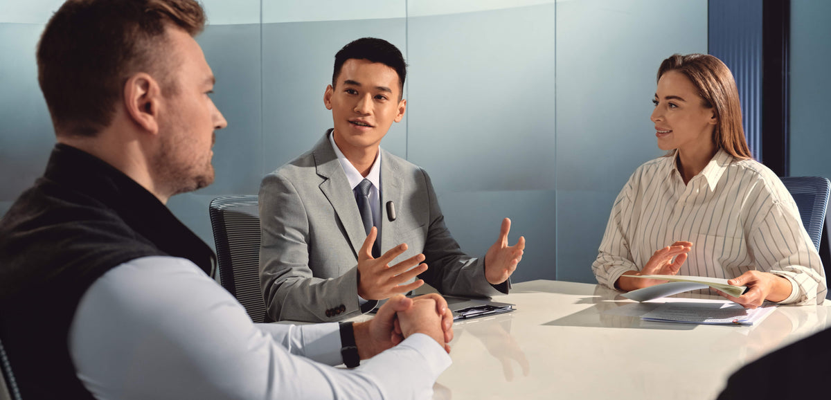 A man wearing a black Plaud NotePin on his suit lapel during a collaborative office meeting.