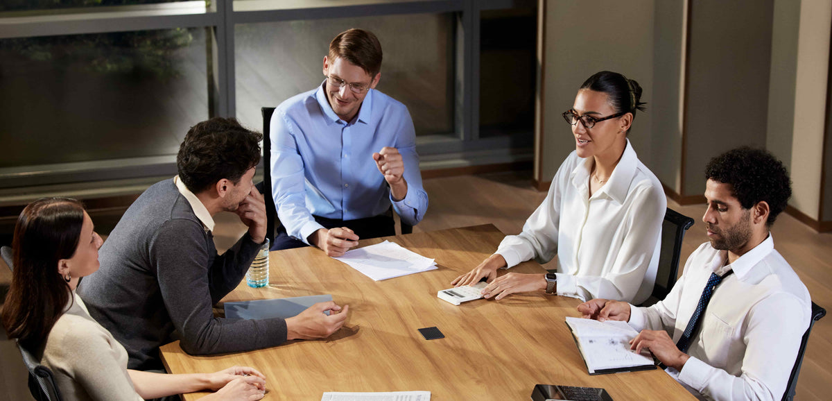 Five professionals in a meeting with a black Plaud Note placed centrally on a wooden conference table.