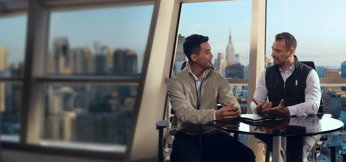 Two men in a city office wearing silver Plaud NotePin devices on a vest and lanyard while talking.