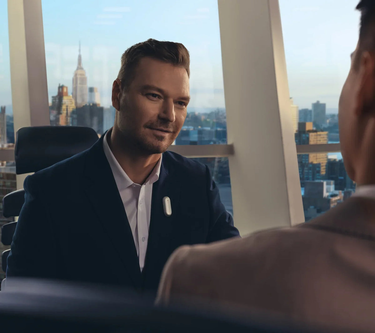 A man in a suit wearing a silver Plaud NotePin on his lapel during a business meeting in a city office.